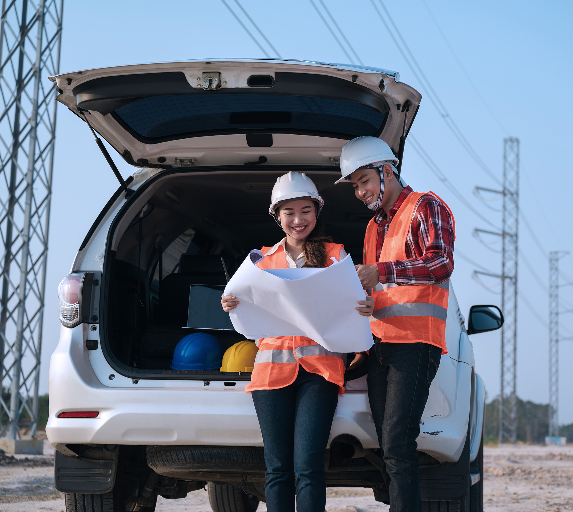 Two construction workers wearing hard hats and orange safety vests looking at blueprints by an open car trunk with helmets inside.