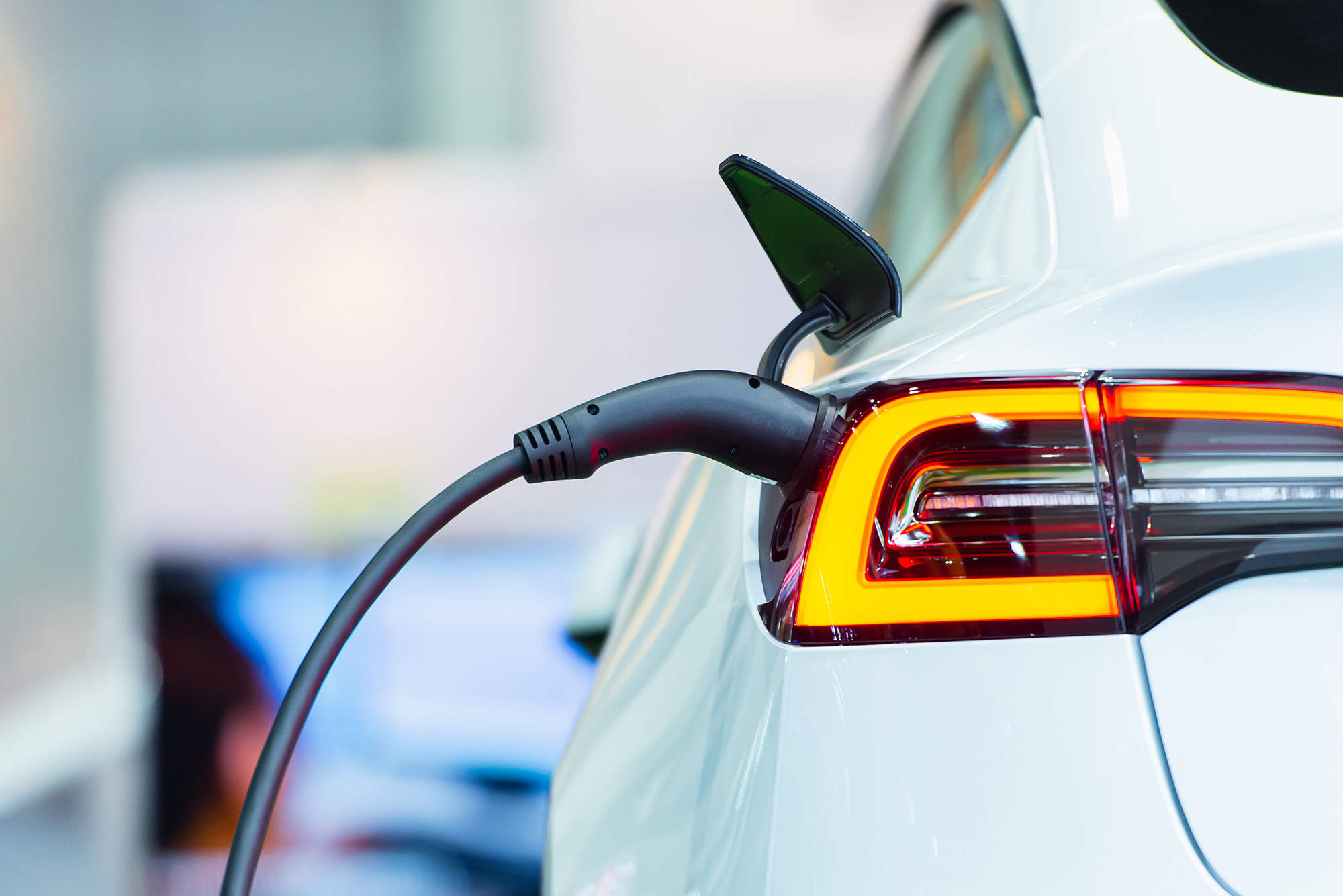 Close-up of a white electric car being charged, showing the charging cable plugged into the rear with illuminated taillight.