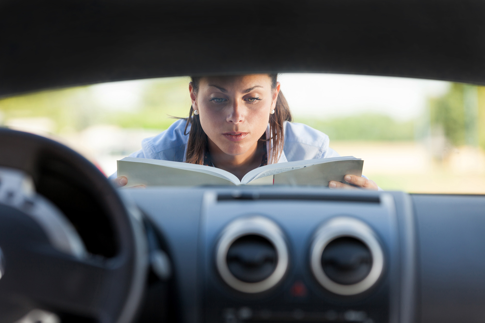 Woman reading a book while leaning over the front of a car, viewed from inside the vehicle.