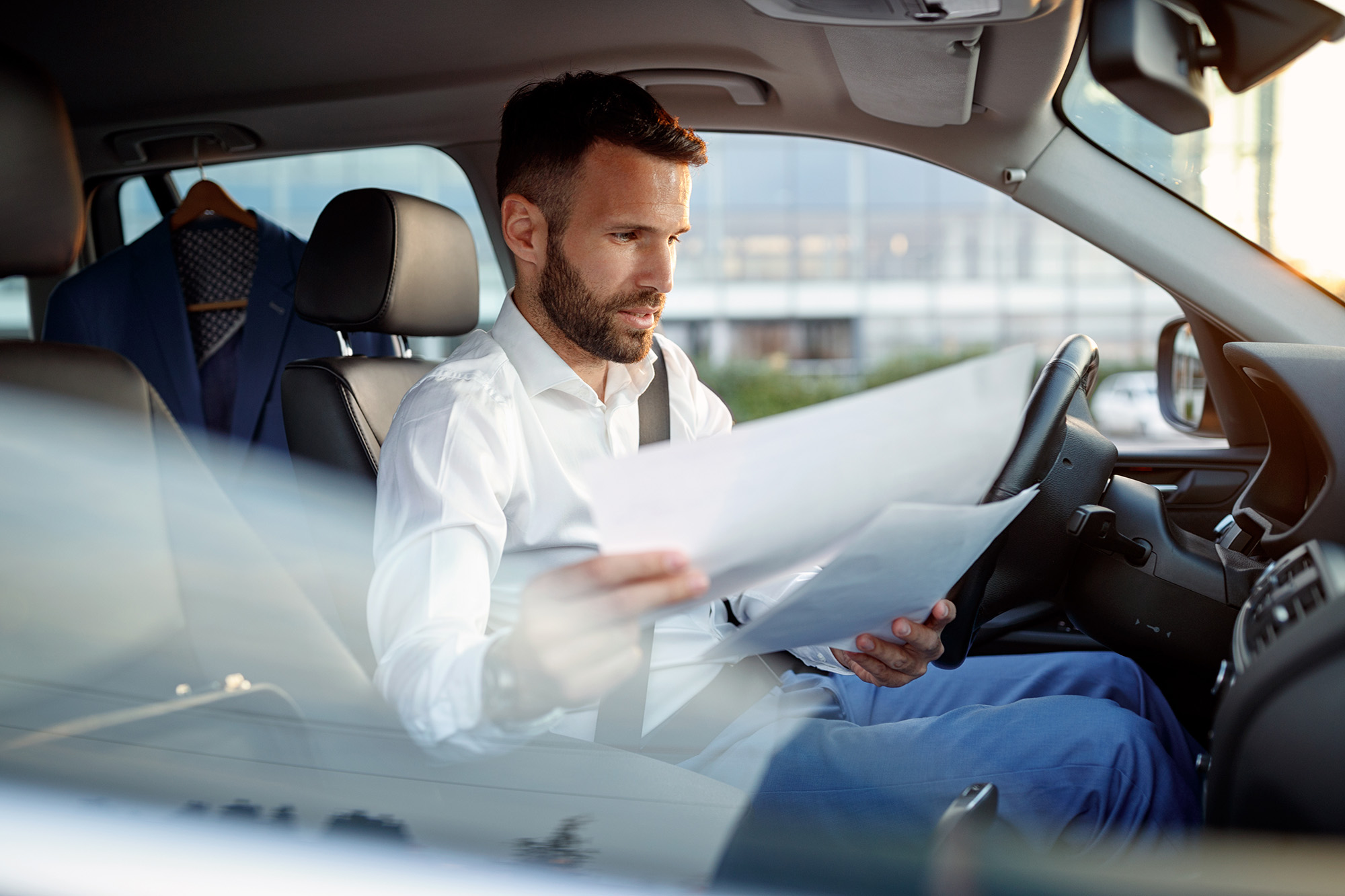 Man in white shirt sitting in a car seat, wearing a seatbelt and reviewing documents.