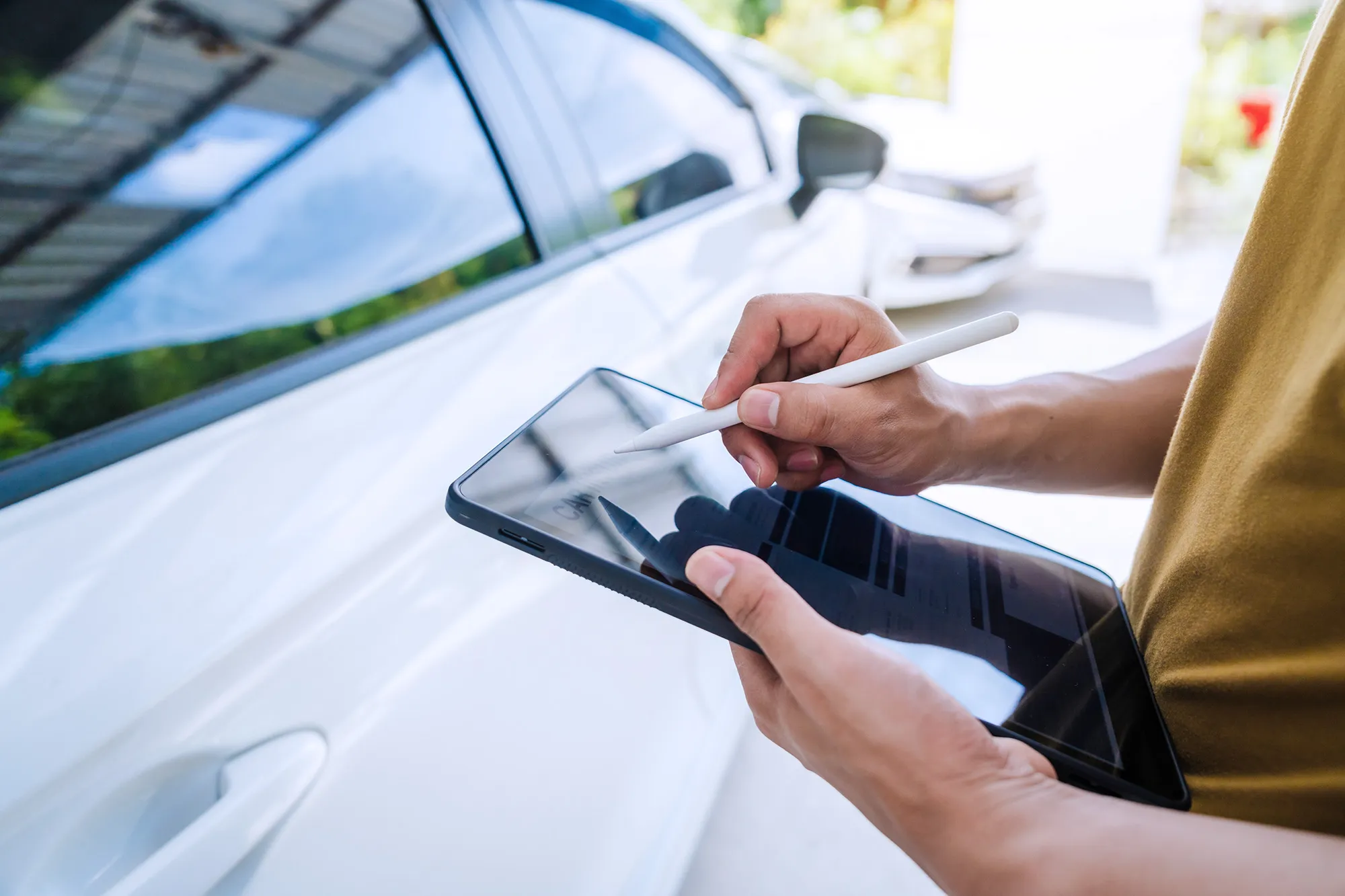 Person holding a tablet and stylus next to a white car, using the tablet for digital notes or inspection.