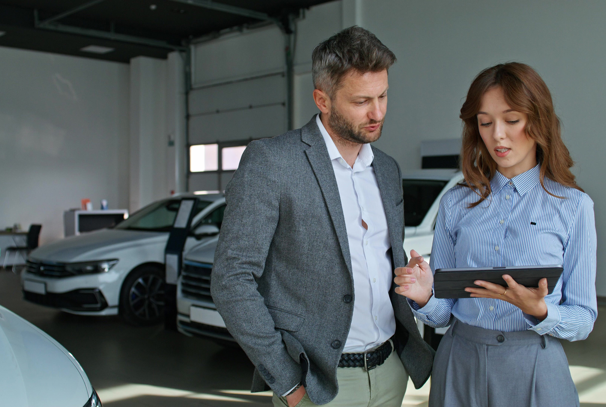 Man in gray blazer and woman in striped shirt discussing and looking at tablet inside a car showroom with white cars in the background.