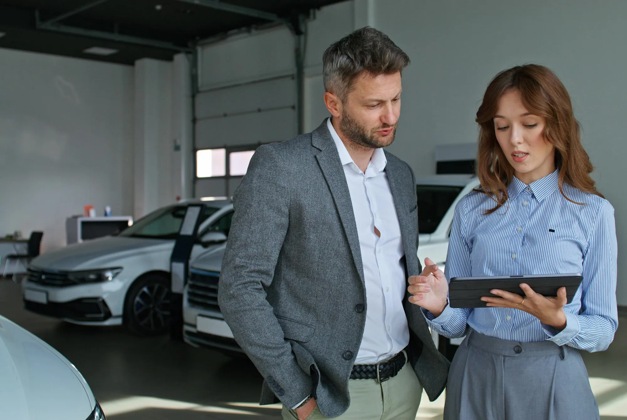Man in gray blazer and woman in striped shirt discussing and looking at tablet inside a car showroom with white cars in the background.