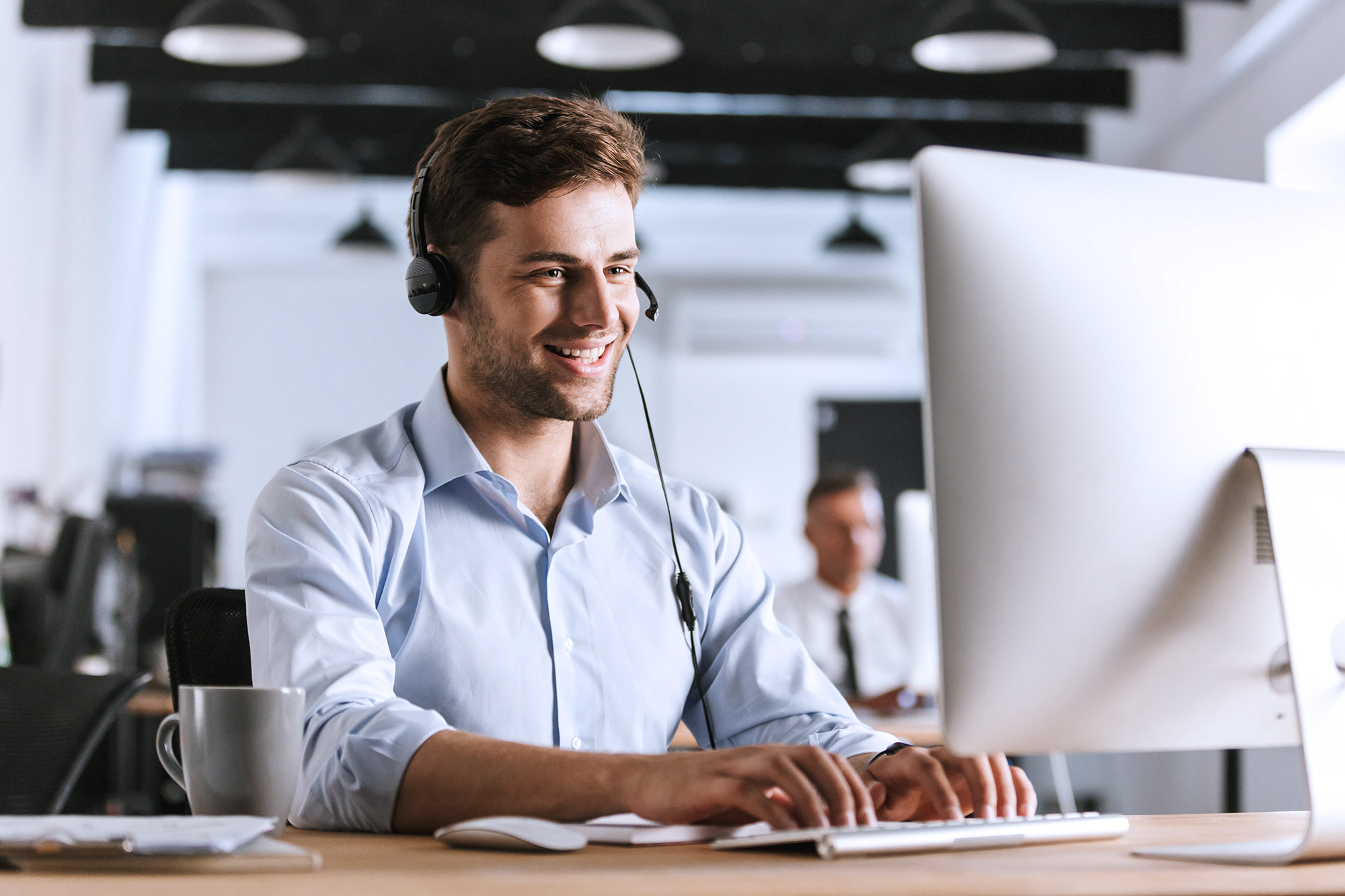 Smiling man wearing a headset and typing on a keyboard in a modern office.