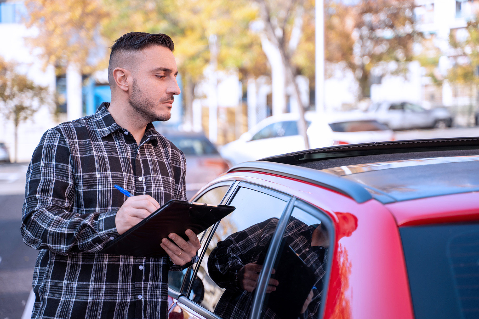 Man in a black and white plaid shirt inspecting a red car and writing on a clipboard outdoors.