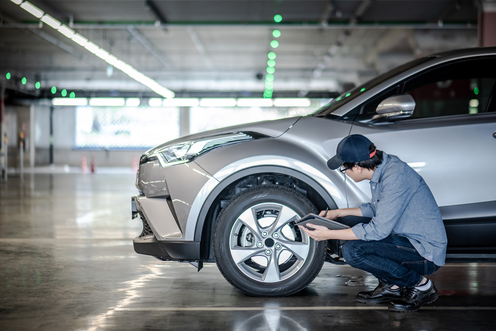 Man wearing a cap and glasses crouching by a silver car's front wheel in a parking garage, inspecting and taking notes on a tablet.