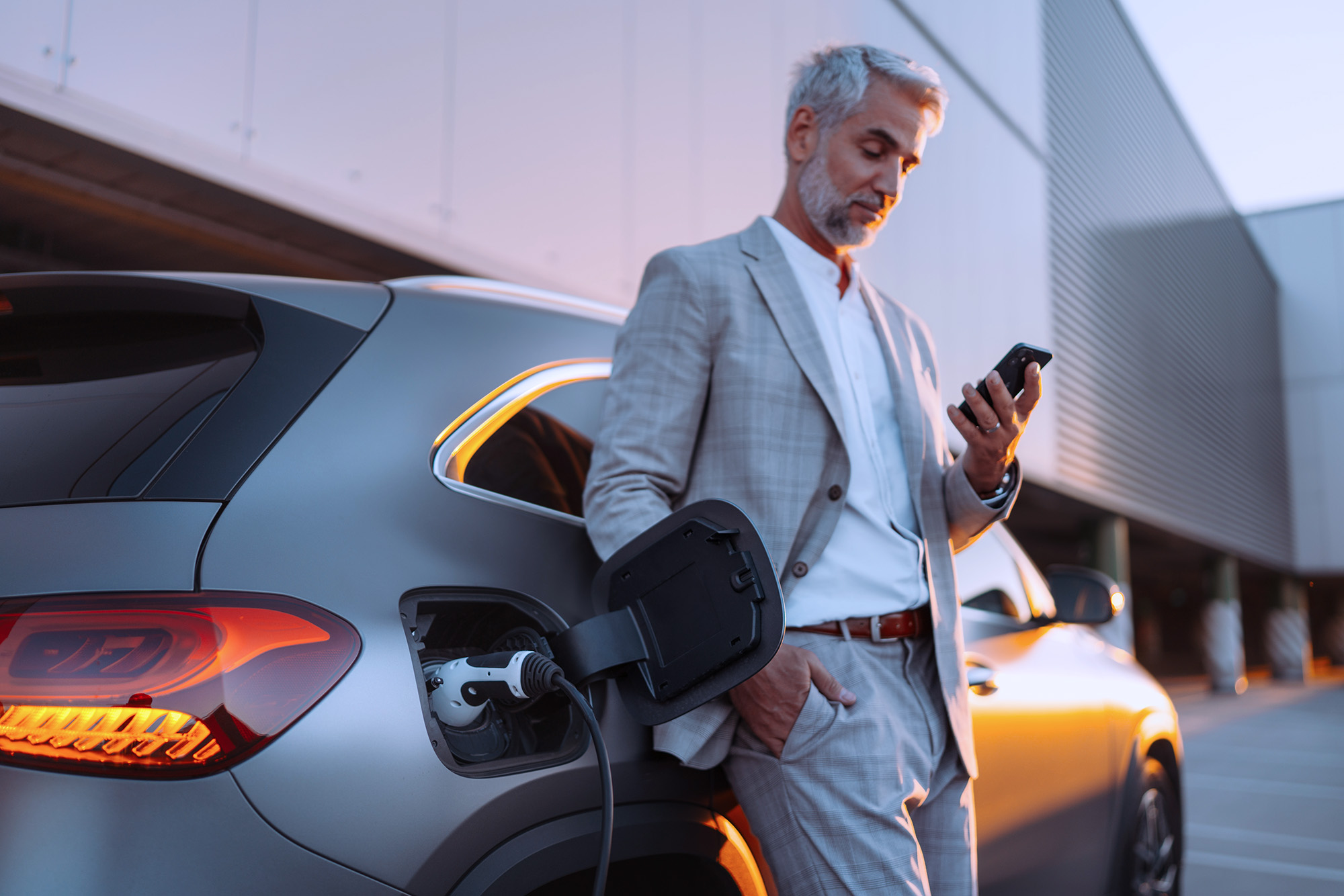 Man in a light gray suit standing next to an electric car charging, looking at his smartphone during sunset.