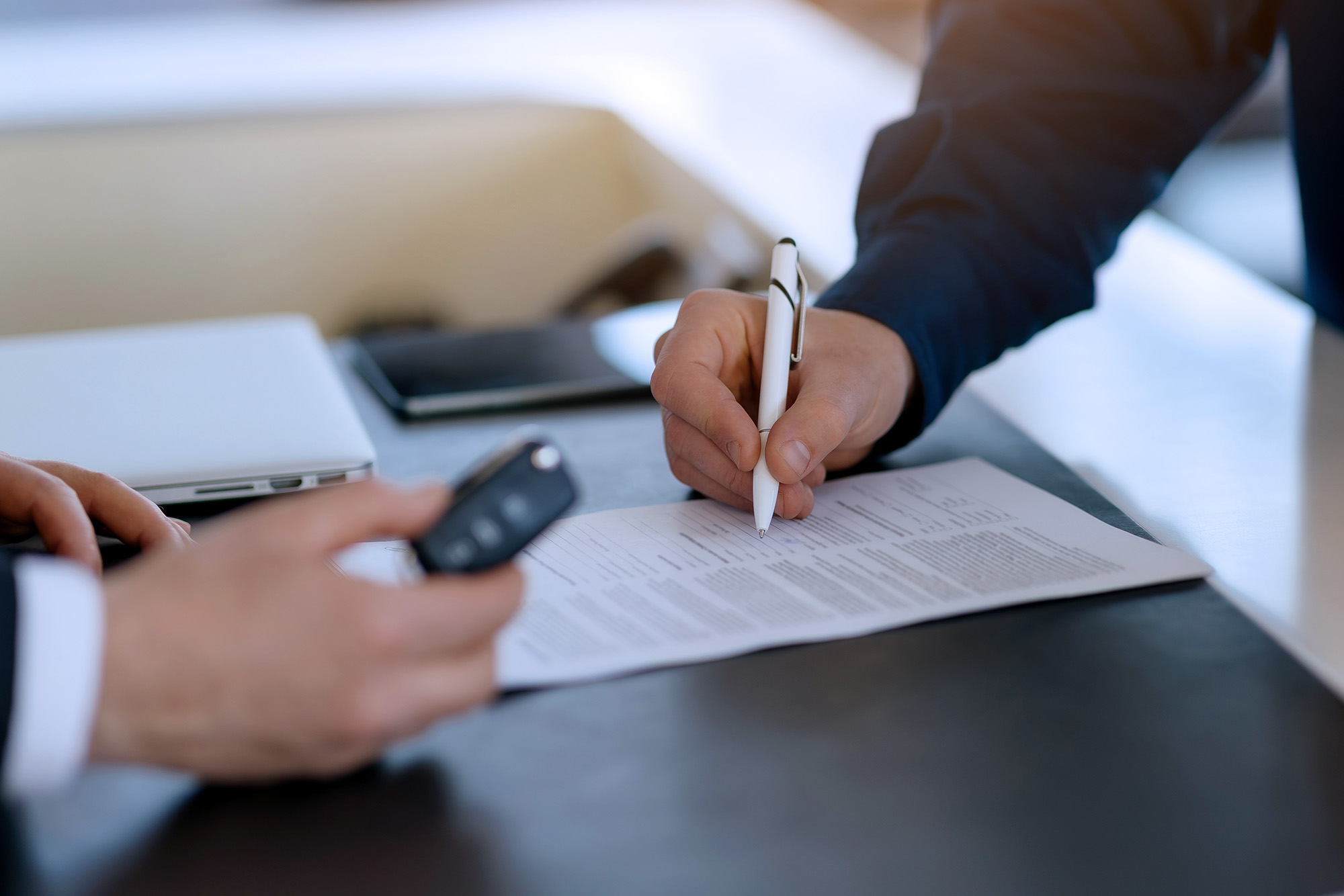 Person signing a document on a desk while another hand holds a car key.