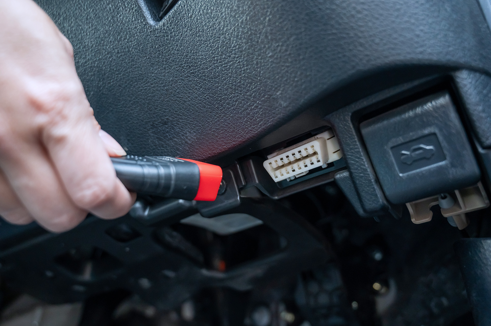 Hand holding a black and red diagnostic tool about to plug into a car's OBD-II port under the dashboard.