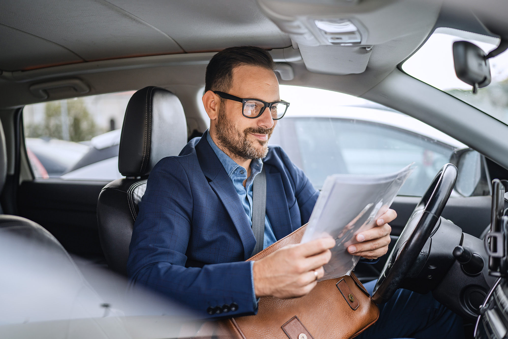 Man in glasses and business attire sitting inside a car, looking at documents from a brown leather folder.