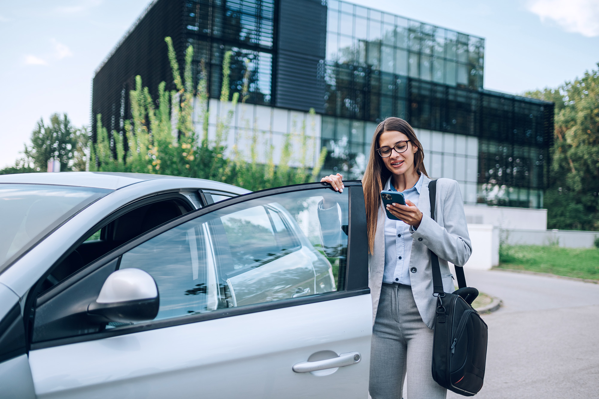 Businesswoman with glasses standing next to an open car door while looking at her phone outside a modern office building.