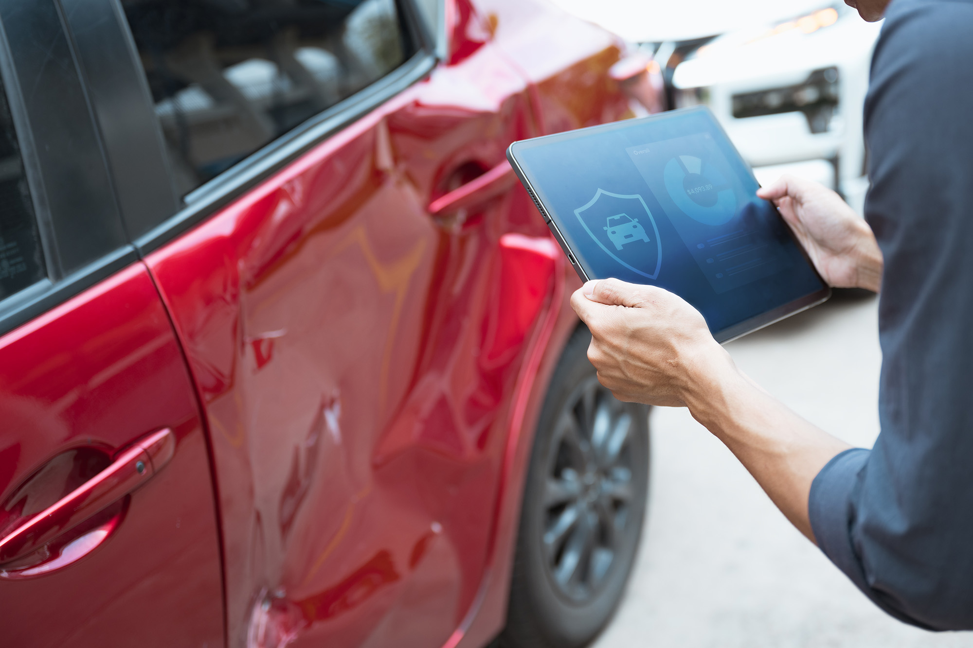 Person inspecting a large dent on the driver's side door of a red car using a tablet with insurance icons visible.