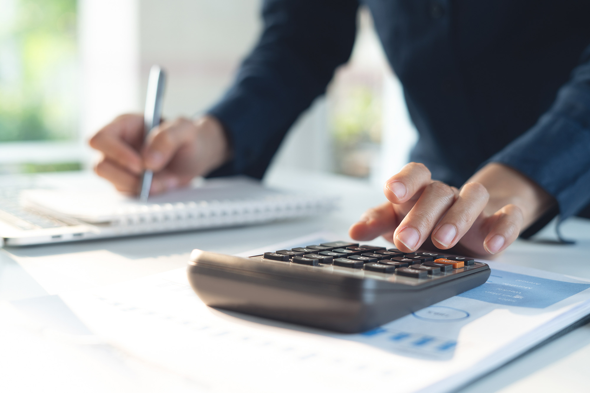 Person using calculator with one hand and writing in a notebook with the other on a desk with documents.