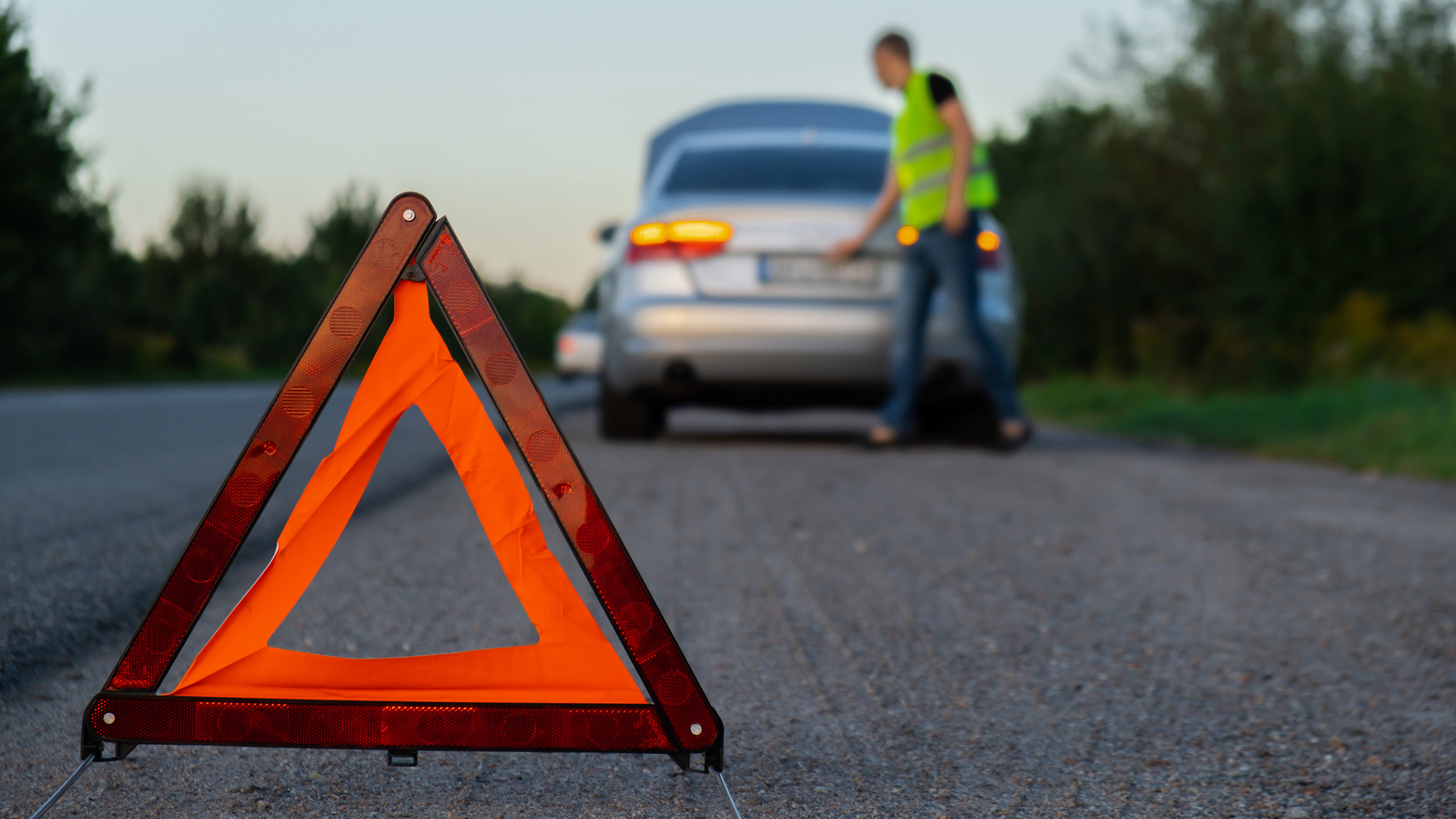 Reflective warning triangle on roadside with a person in a high-visibility vest near a silver car with open trunk in the background.