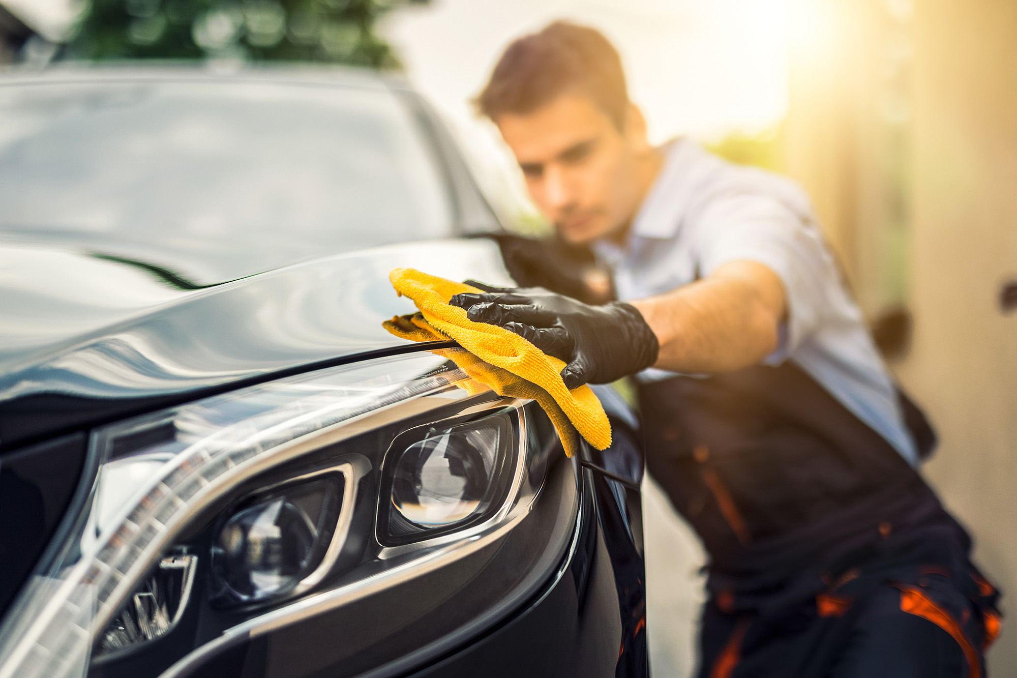 Person wearing black gloves polishing a black car's headlight with a yellow cloth.