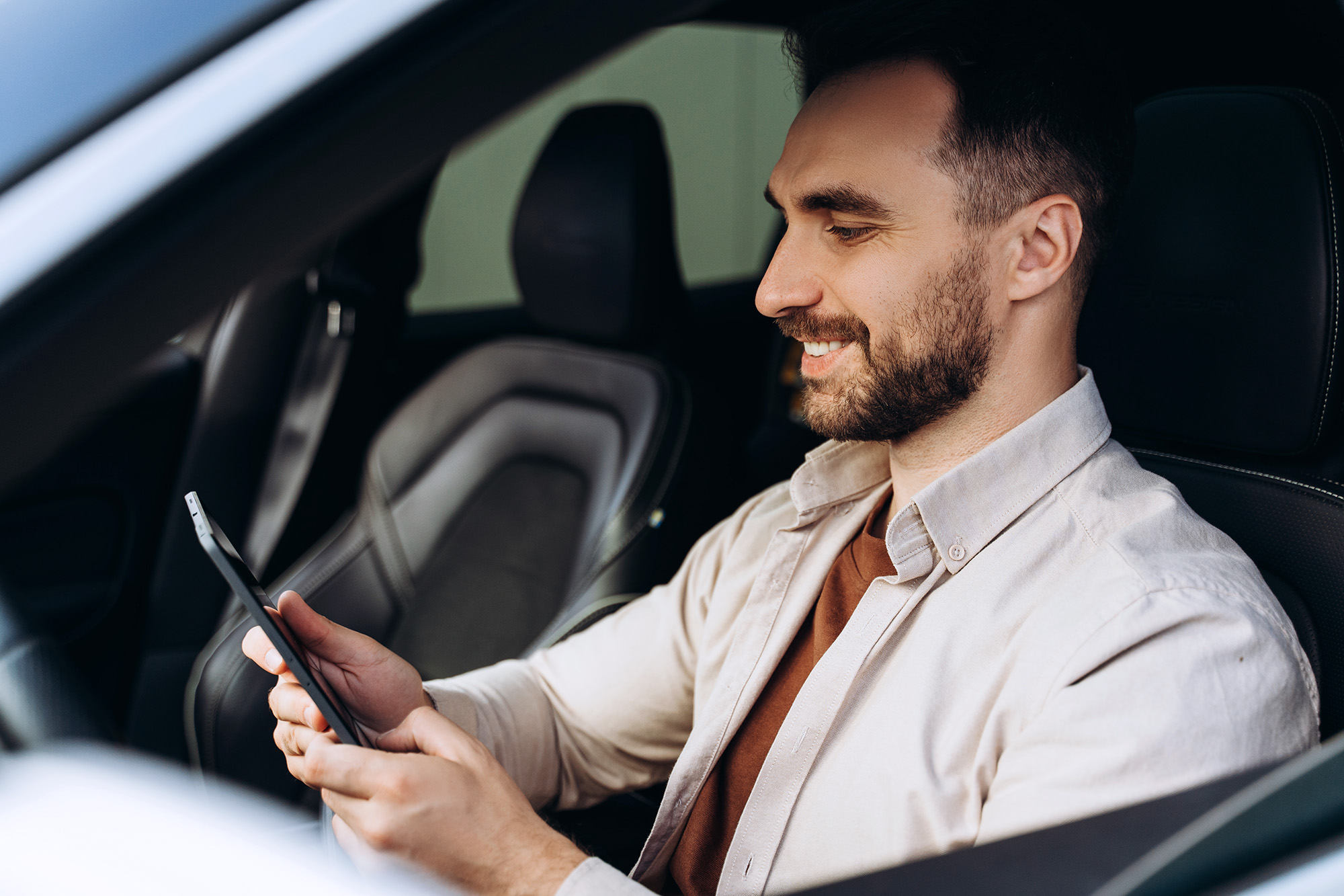 Smiling man sitting in car holding and looking at a tablet device.