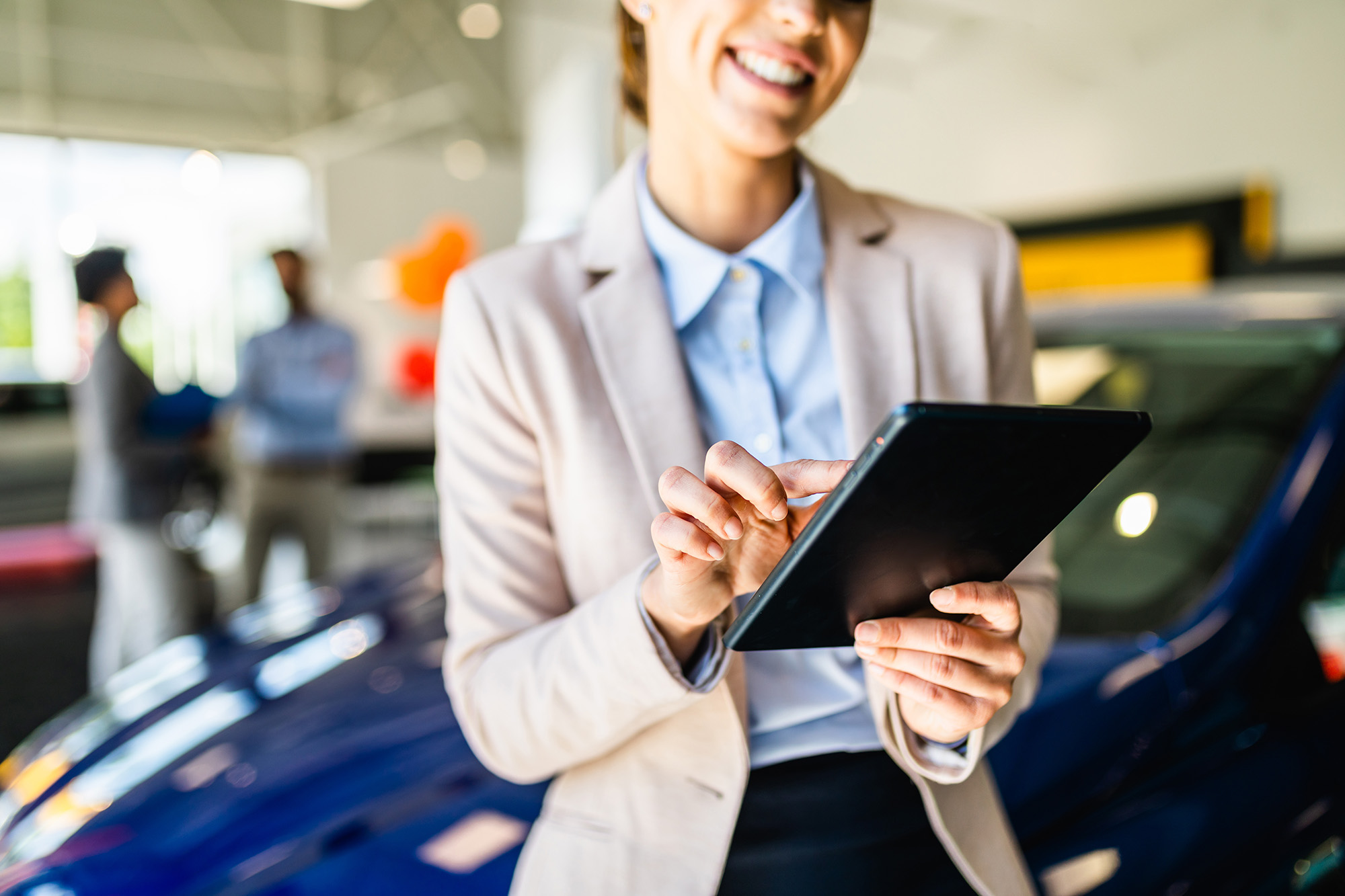 Smiling woman in a beige jacket using a tablet in a car showroom with a blue car and two blurred people in the background.