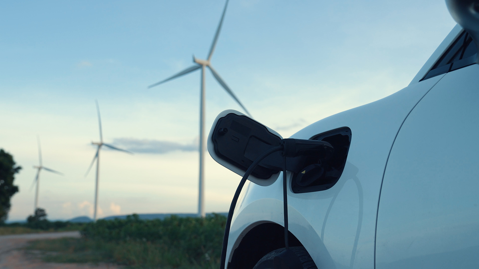 Close-up of a white electric car plugged in for charging with several wind turbines in the background under a clear sky.