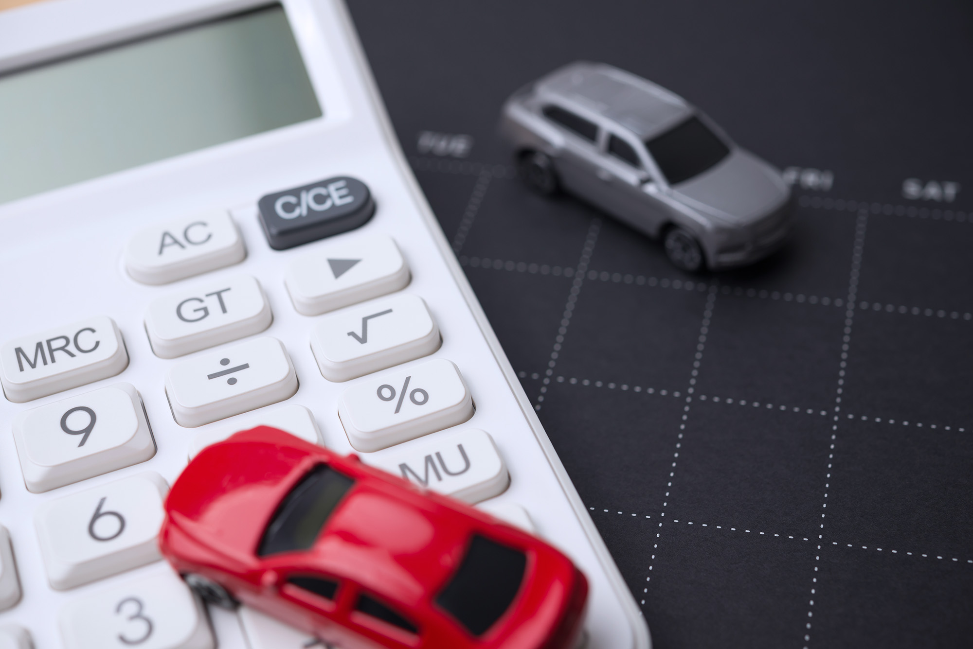Red toy car on white calculator and gray toy car on black calendar with weekdays.
