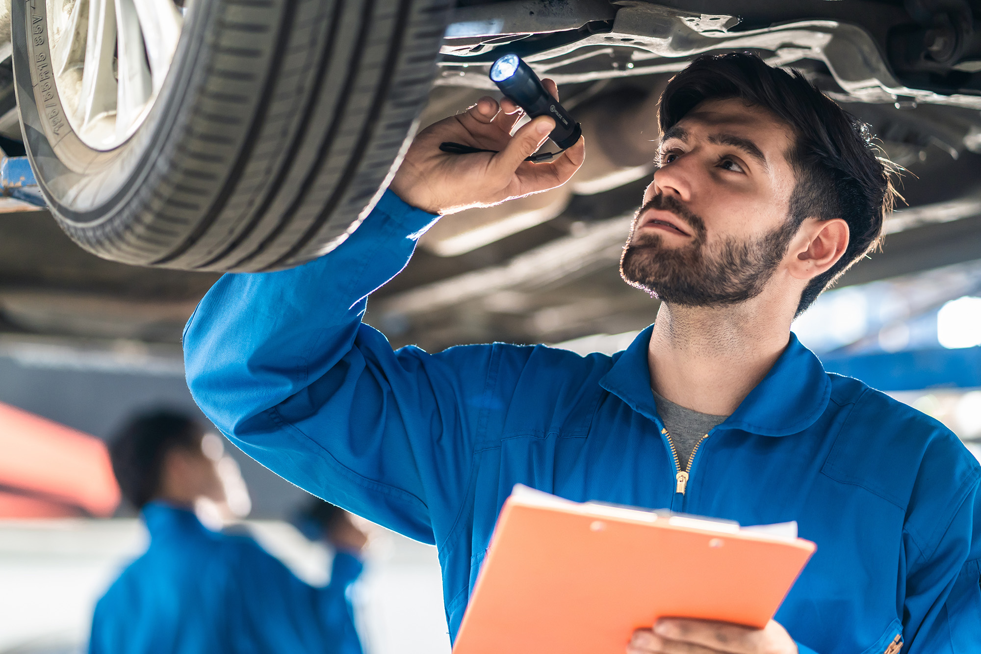 Mechanic in blue coveralls inspecting the underside of a vehicle with a flashlight while holding a clipboard.