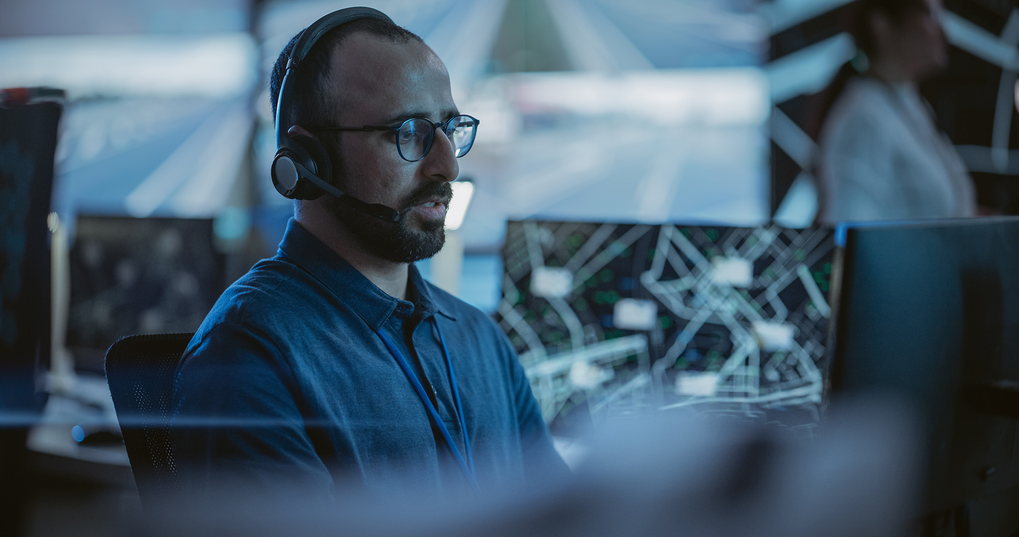 Man wearing glasses and a headset sitting in front of multiple computer monitors displaying maps and data in a dimly lit control room.