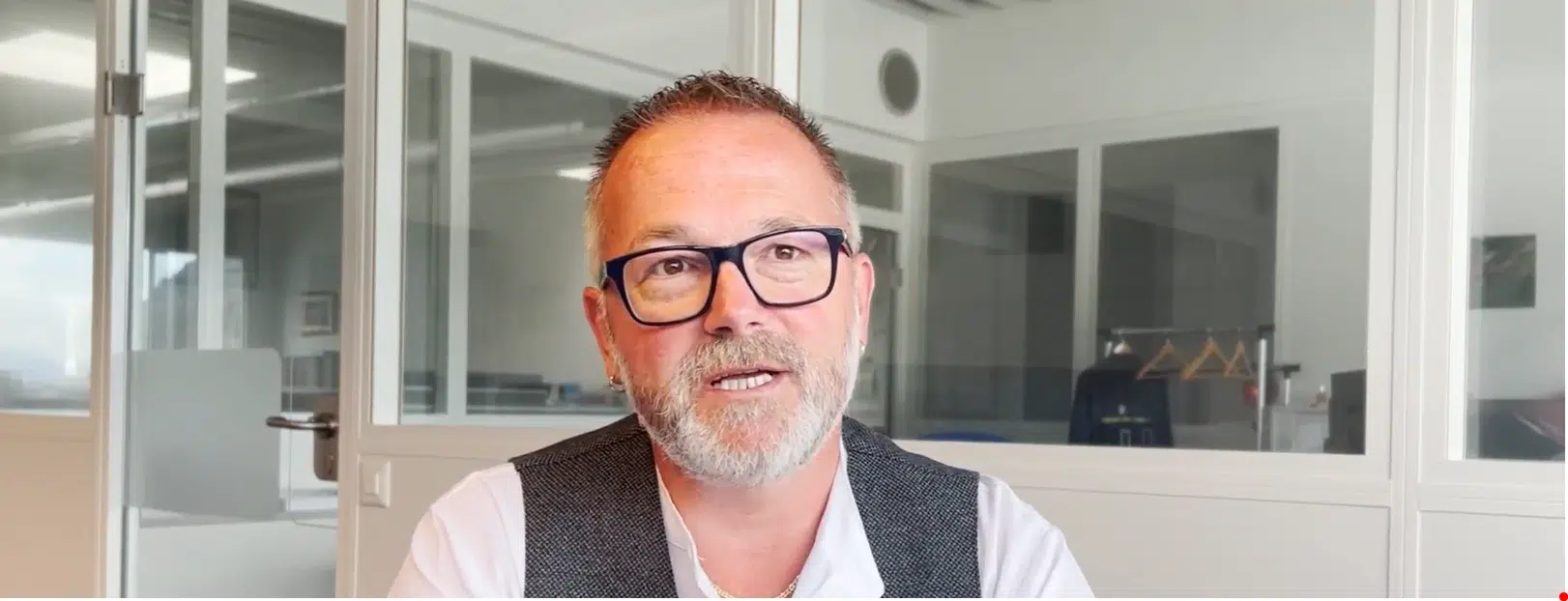 Middle-aged man with short hair, beard, glasses, and white shirt with a dark vest sitting in a modern office with glass partitions.