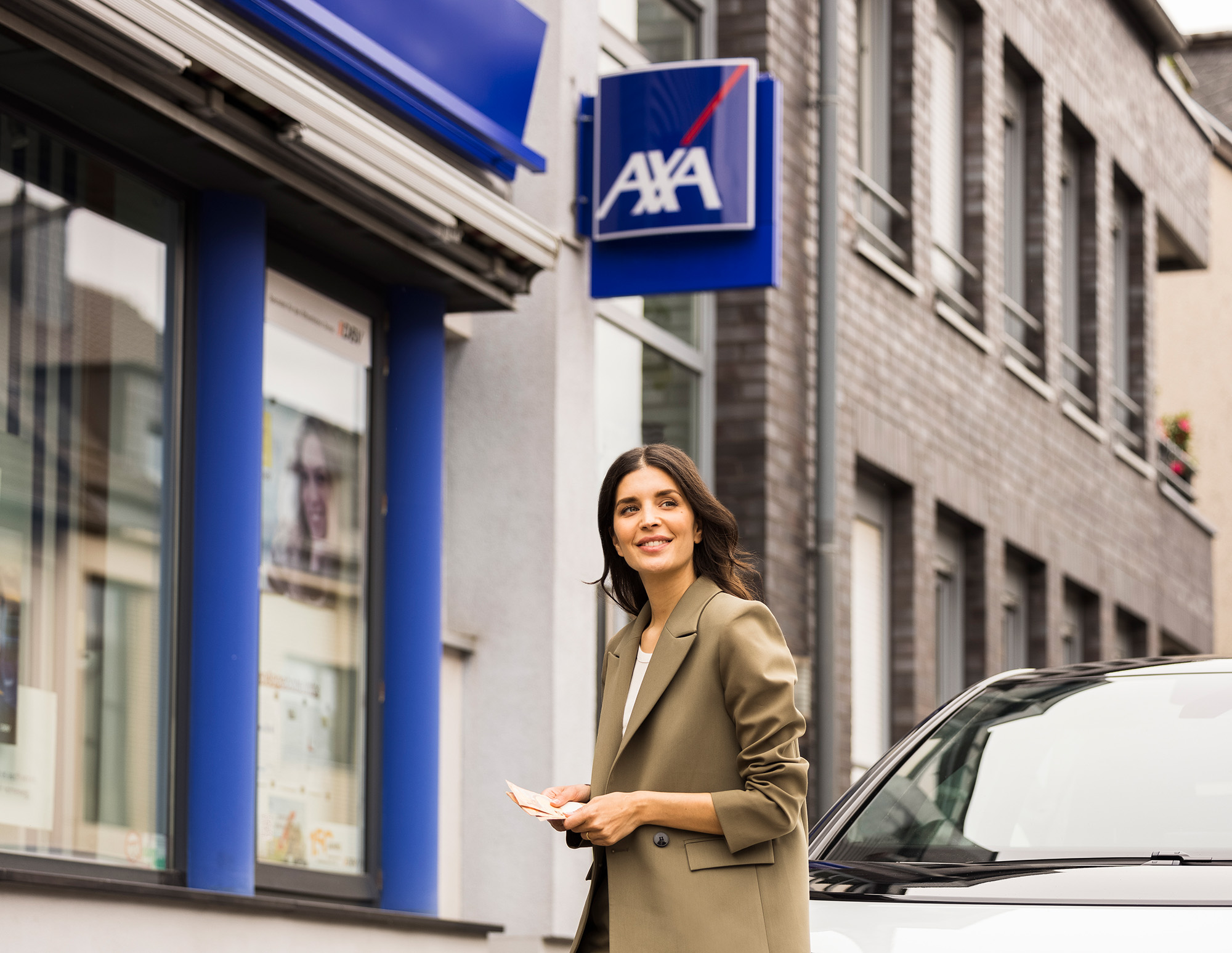 Smiling woman holding banknotes standing beside a white car in front of an AXA insurance office.
