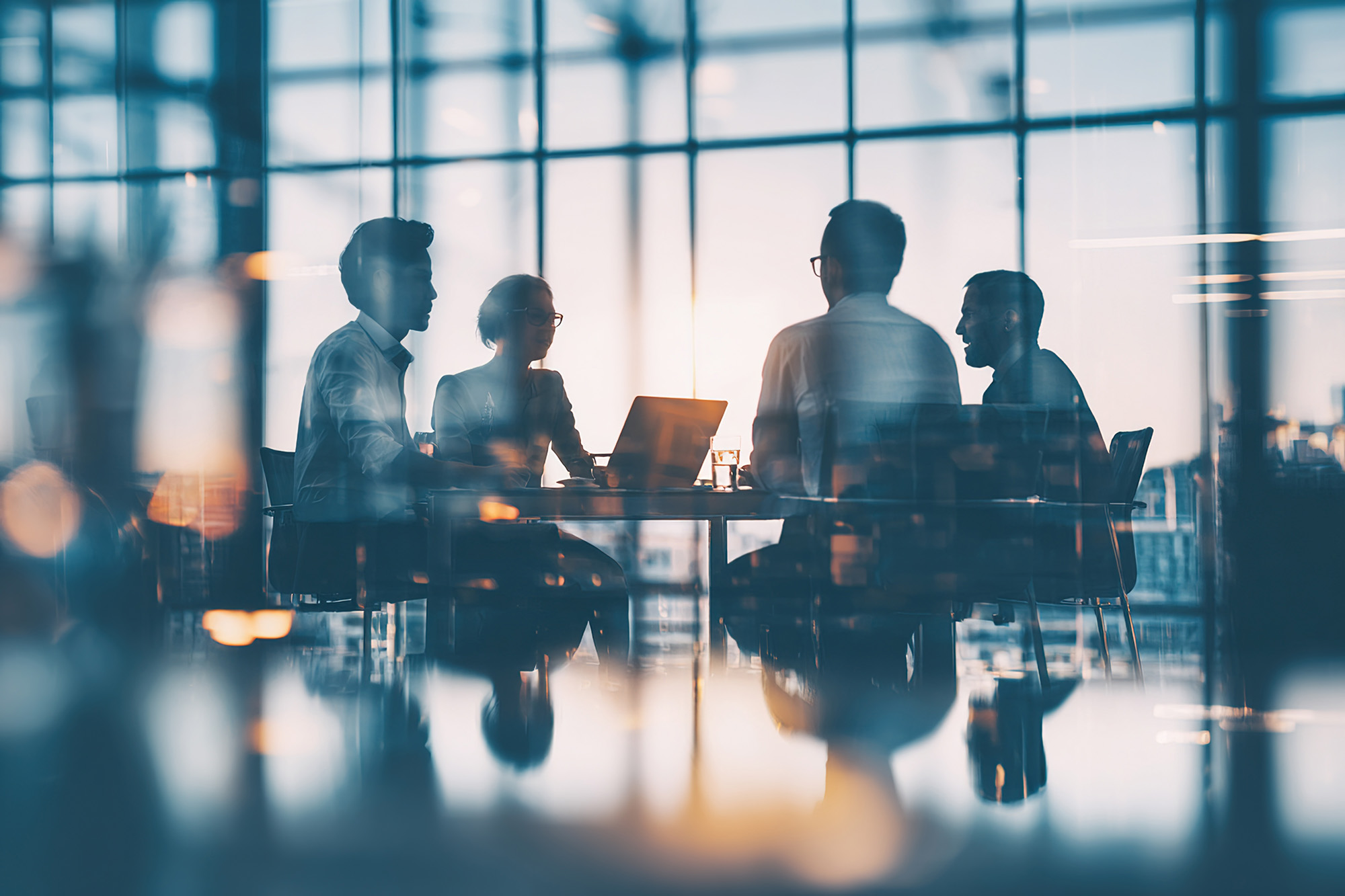 Four businesspeople sitting around a table engaged in a meeting with laptop in a modern office with large windows.