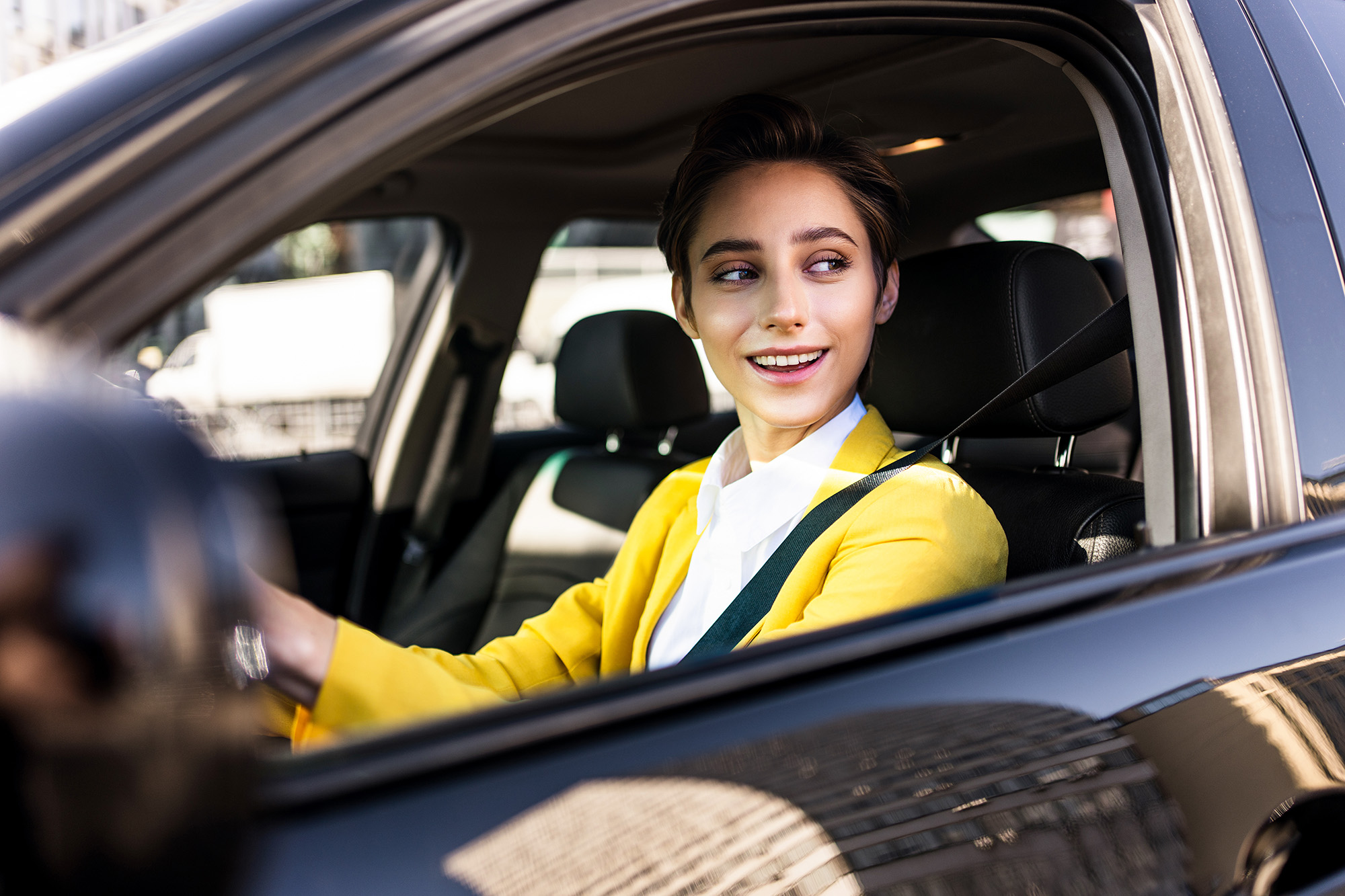 Smiling young woman wearing a yellow blazer and seatbelt driving a black car.