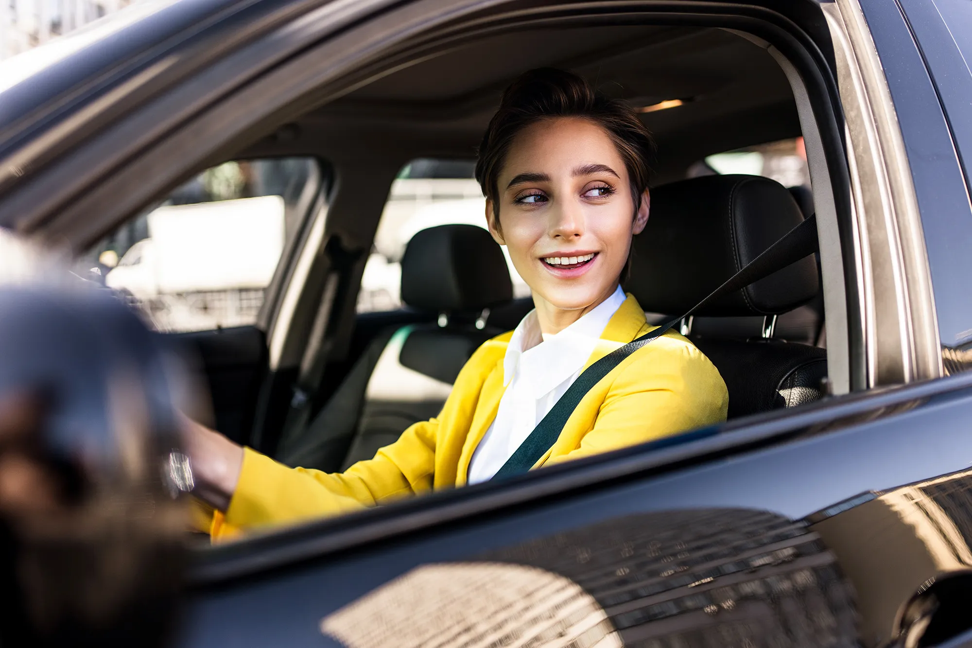 Smiling young woman wearing a yellow blazer and seatbelt driving a black car.