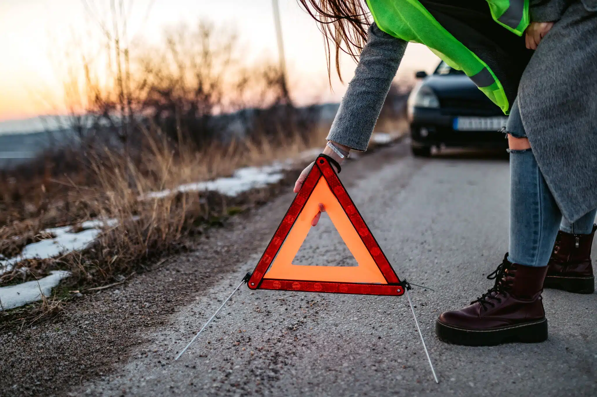 Person wearing a reflective safety vest placing a red warning triangle on a road near a parked black car at dusk.