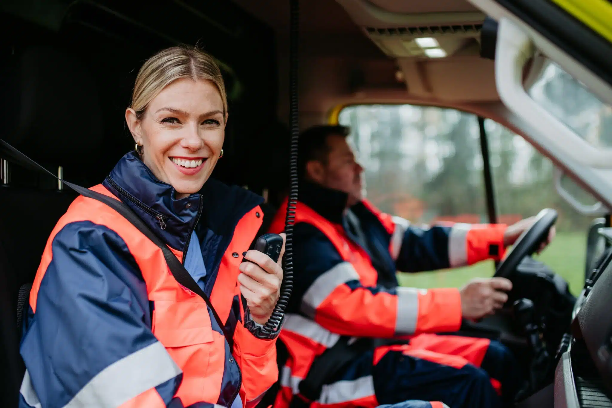 Two emergency responders wearing orange and navy reflective jackets sitting inside an ambulance, one smiling and holding a radio while the other drives.