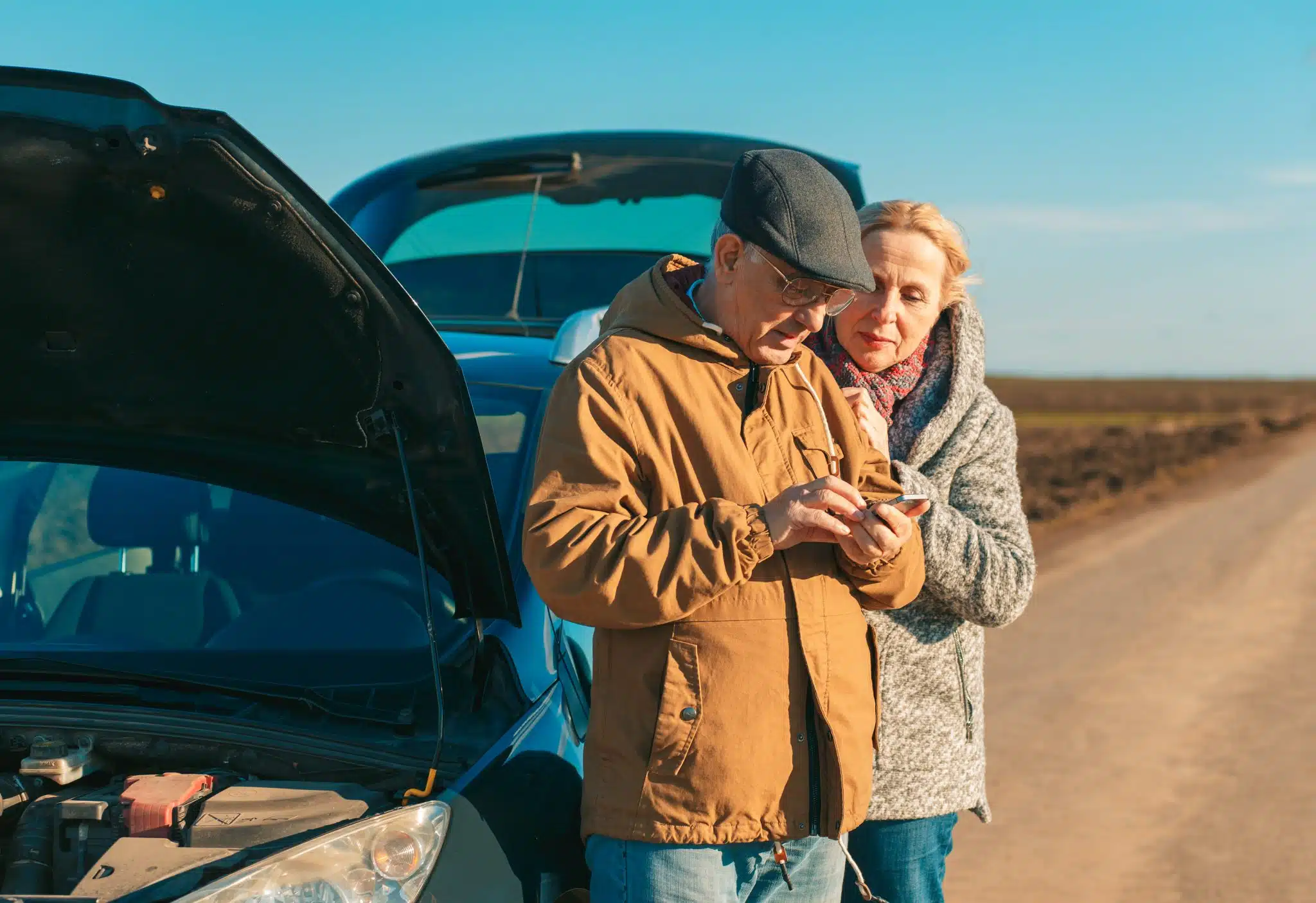 An elderly couple stands beside a broken-down car with an open hood on a rural road, looking at a smartphone.