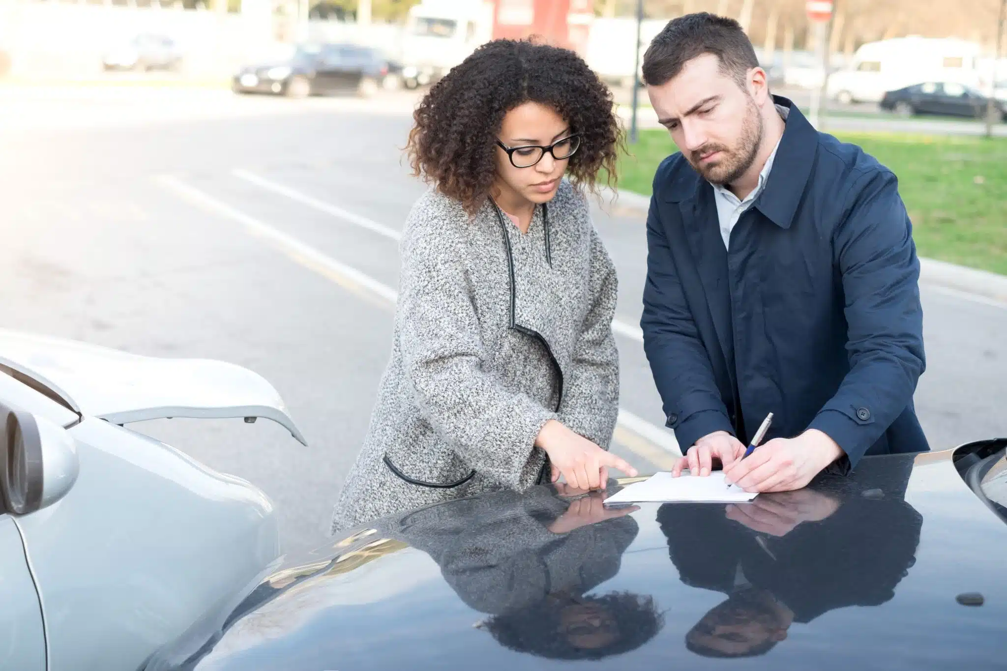Two adults reviewing and signing documents on the hood of a car after a minor traffic accident.