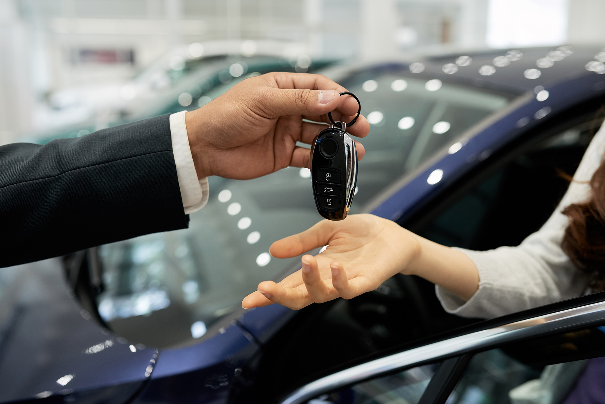 Person handing over a car key fob to another person sitting inside a blue car.
