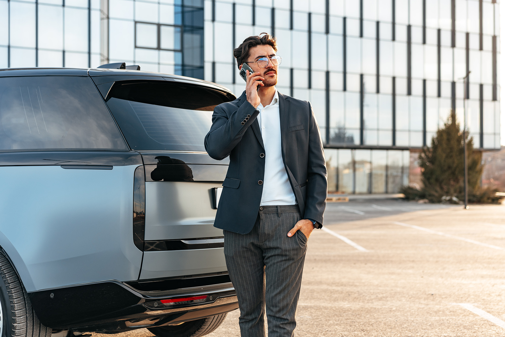 Man in a dark blazer and striped pants standing beside a modern silver SUV while talking on a smartphone in a parking lot.