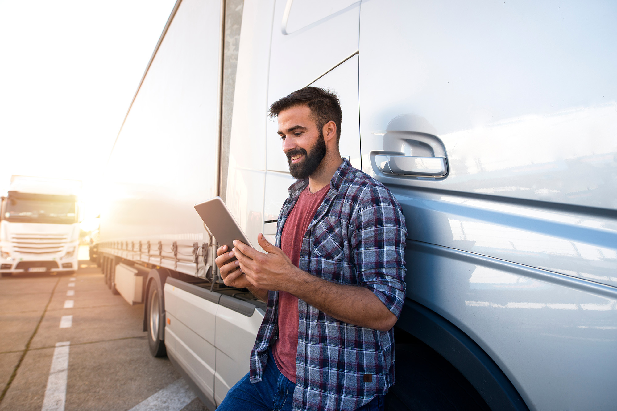 Smiling man with beard using a tablet while leaning against a white semi-truck trailer on a sunny day.