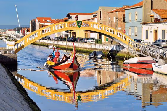 Traditional boat with people rowing under a yellow arched bridge over a calm canal in a town.