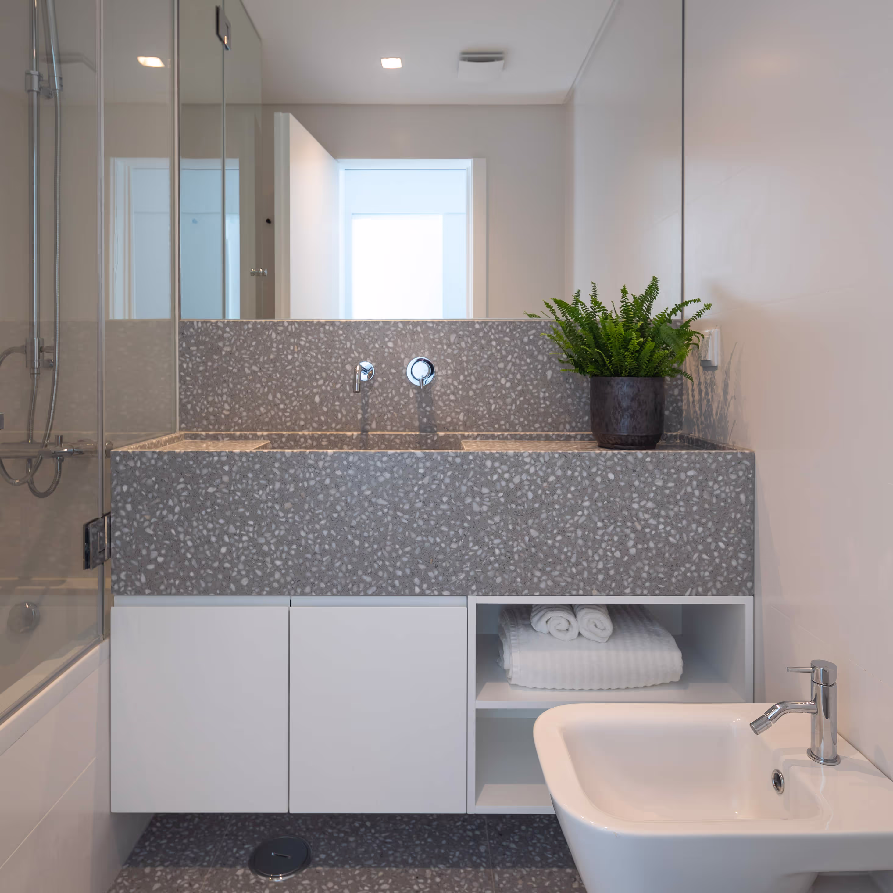 Modern bathroom sink with terrazzo countertop, white cabinetry, folded towels, and a potted green plant.