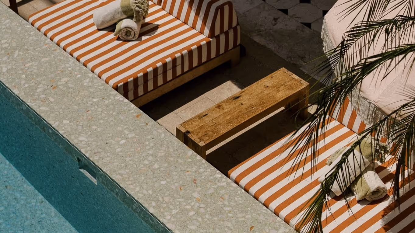 Outdoor seating area with orange and white striped cushions beside a turquoise swimming pool, featuring rolled towels and a wooden table.