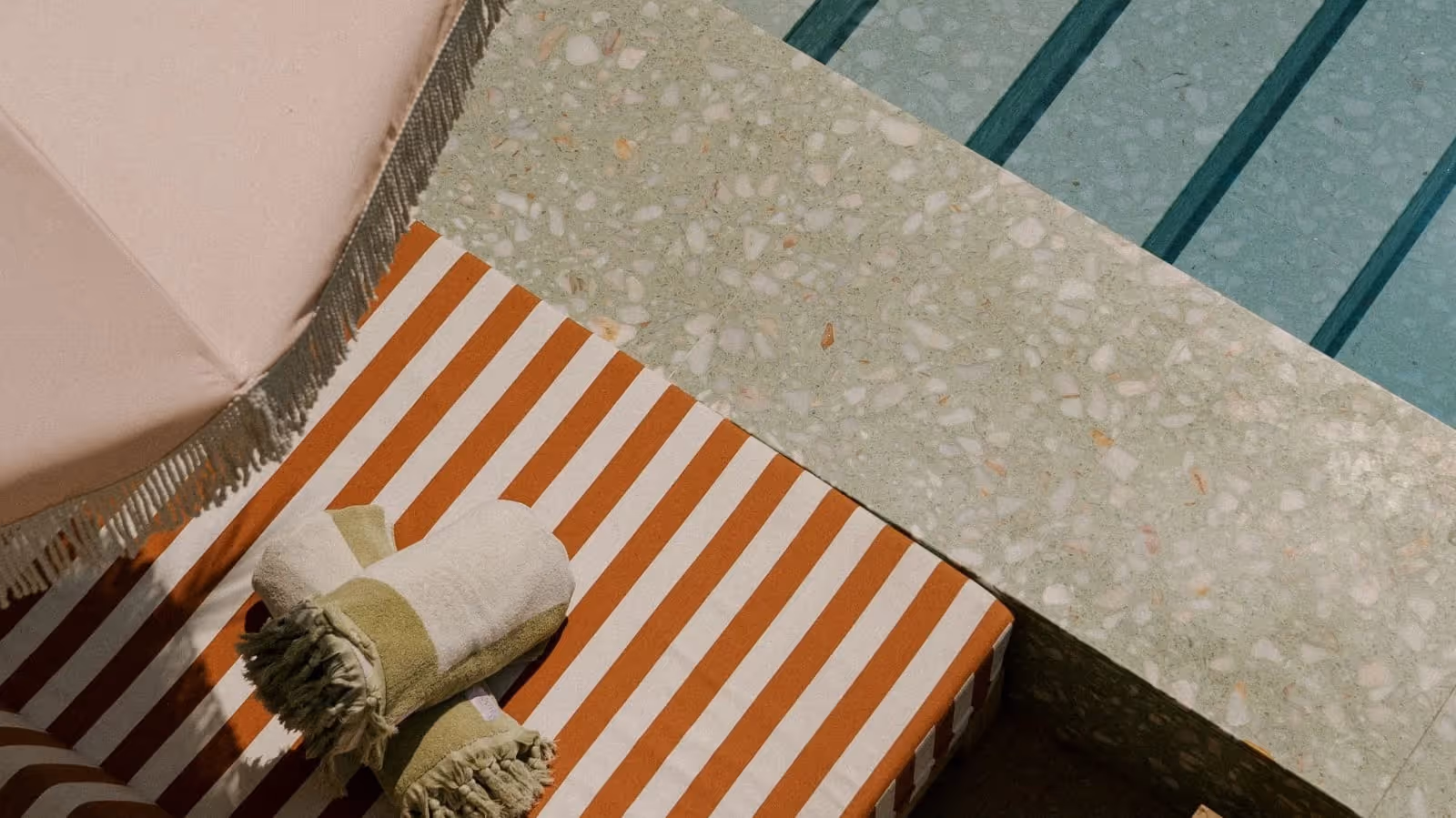 Poolside scene with orange and white striped cushion, two rolled towels, terrazzo pool edge, and turquoise water with dark stripes.