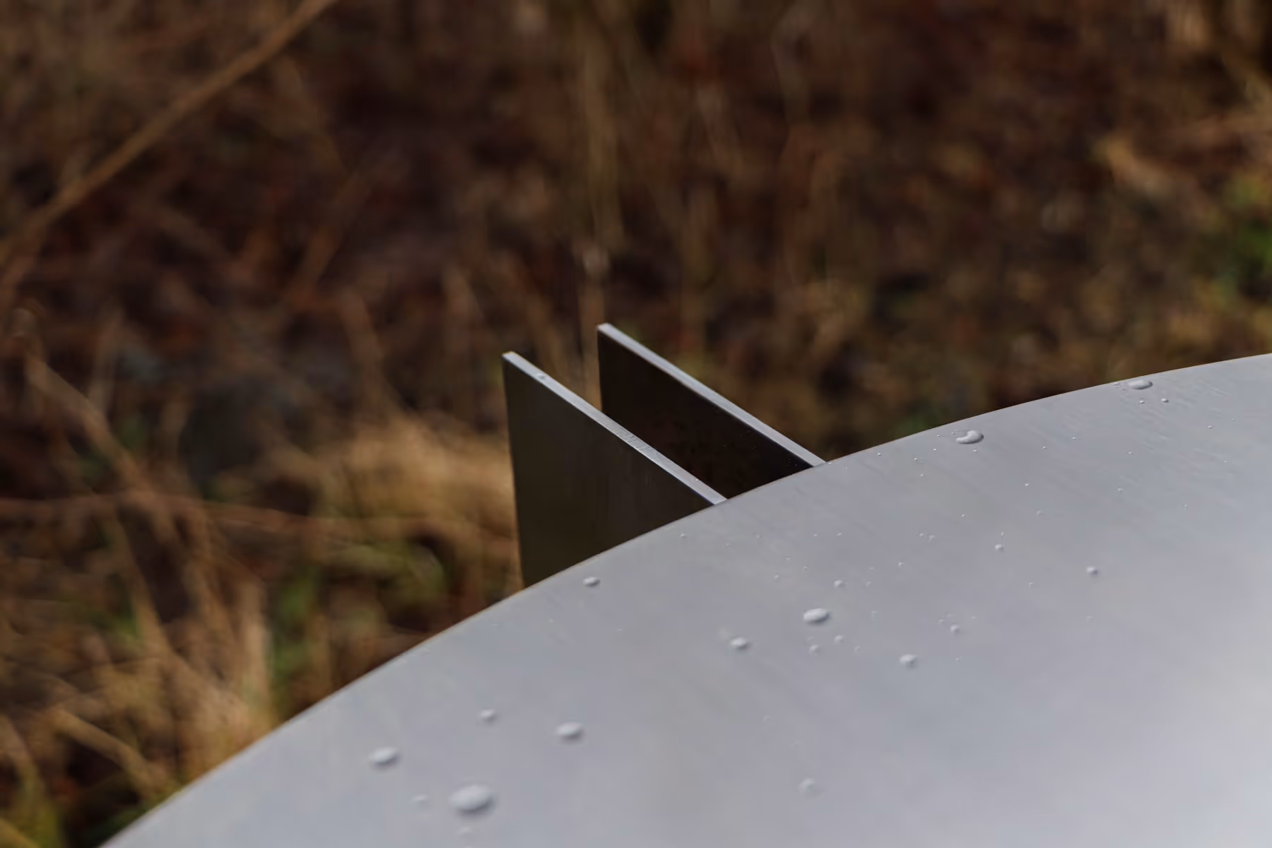 Close-up of a metallic surfboard fin with water droplets on its surface and a blurred natural background.