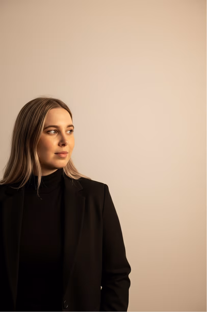 Young woman with long blonde hair wearing a black blazer and turtleneck looking to her left against a beige background.