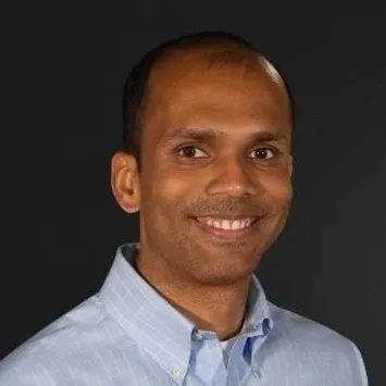 Man with dark skin and short hair smiling, wearing a light blue button-up shirt against a black background.