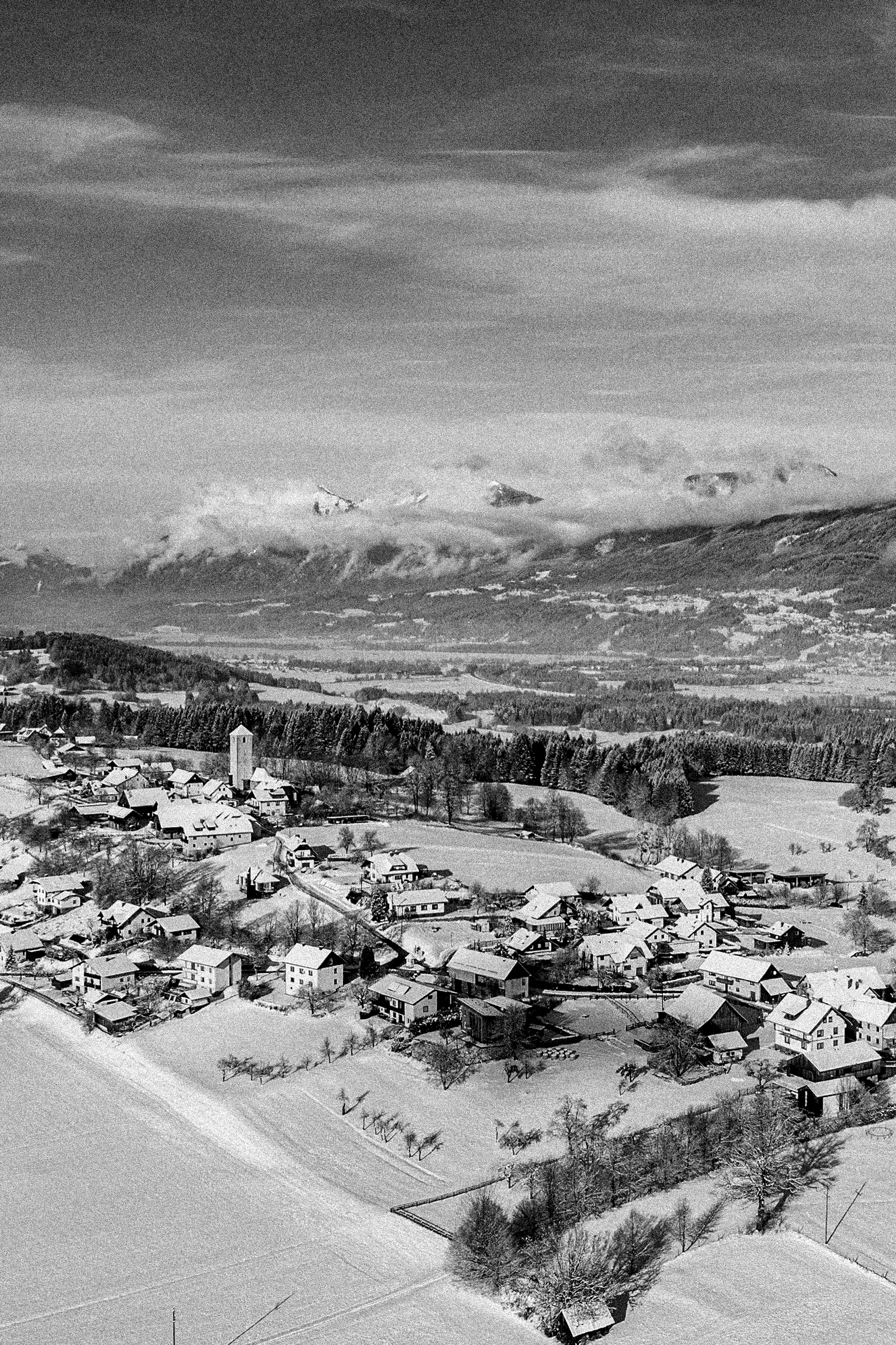 Schneebedecktes Dorf mit Häusern, Feldern und Bäumen vor einer Bergkette mit Wolken.