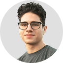 Headshot of a young man with curly hair and black glasses wearing a dark shirt, smiling slightly against a white background.