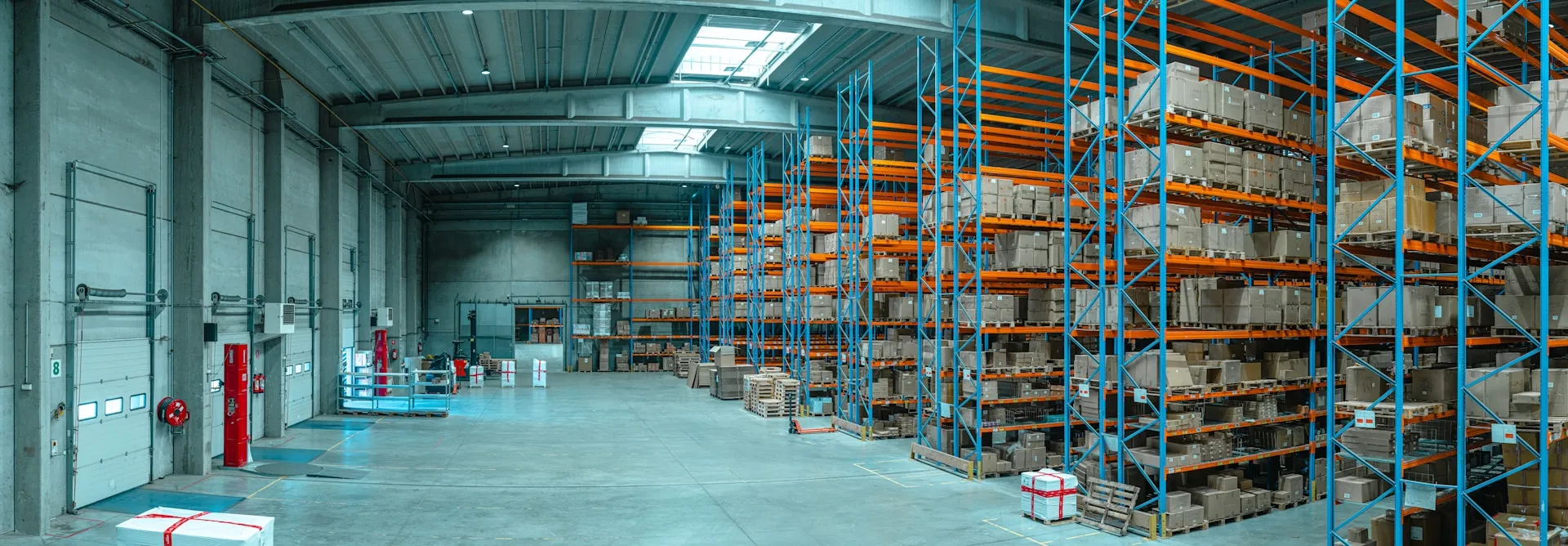 Spacious warehouse with tall blue and orange racks filled with stacked cardboard boxes and pallets on a concrete floor.