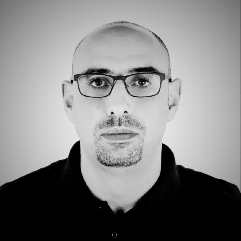 Black and white portrait of a bald man with glasses and a goatee, wearing a dark collared shirt.