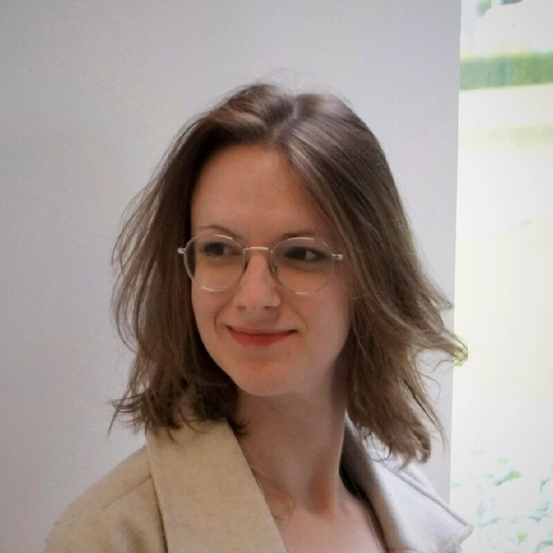 Smiling woman with shoulder-length brown hair and glasses wearing a beige coat looking to her right.