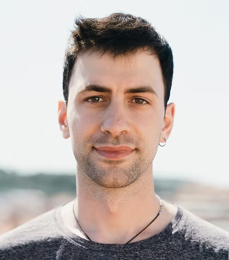 Young man with short dark hair, light beard, and a small hoop earring, wearing a gray shirt and a necklace, standing outdoors with a blurred background.